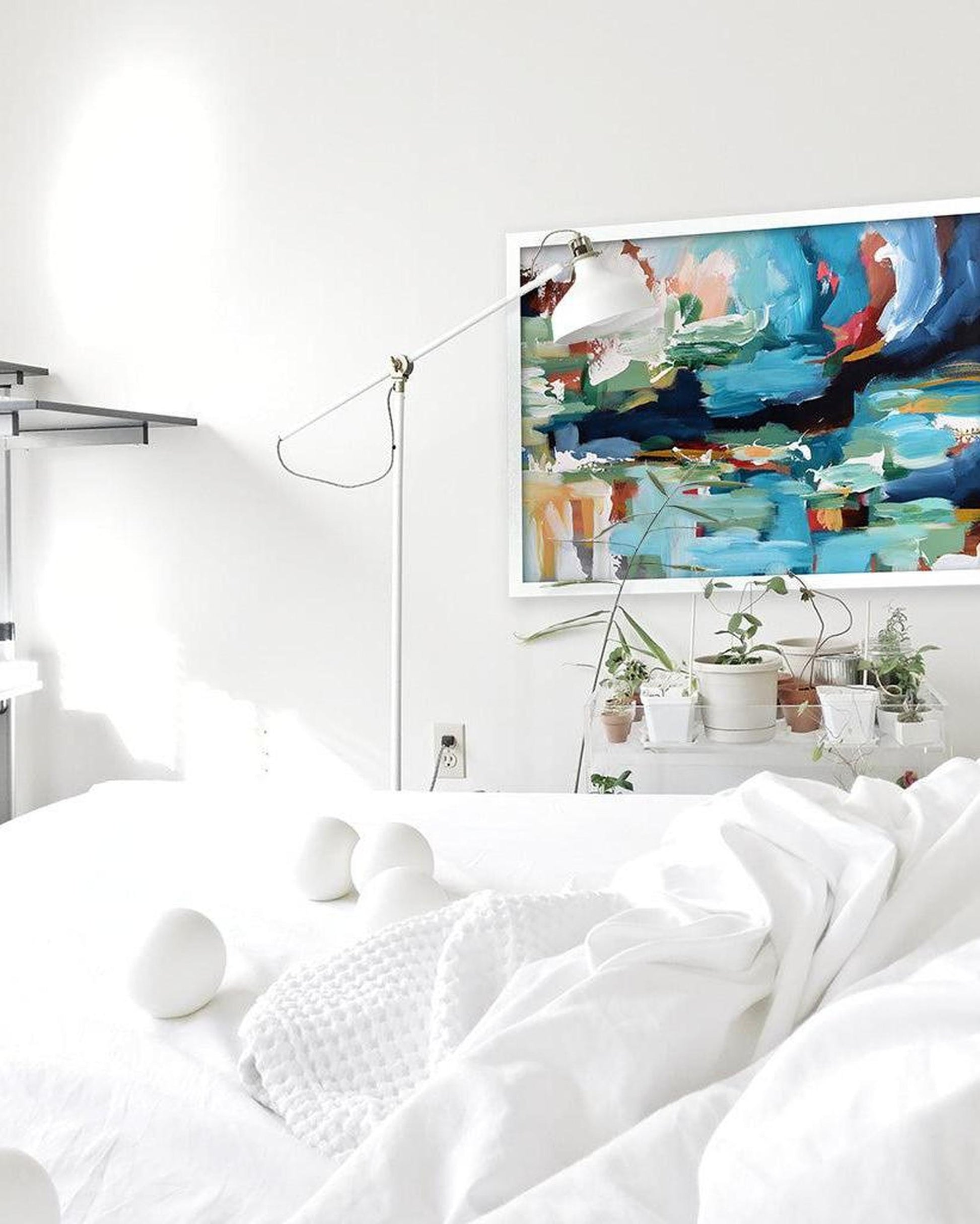 Modern bedroom with white bedding, abstract painting on the wall, and decorative plants.