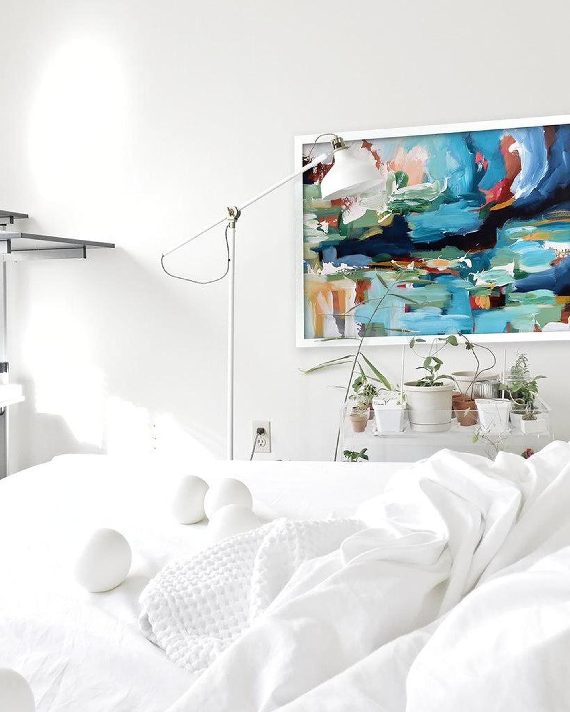 Modern bedroom with white bedding, abstract painting on the wall, and decorative plants.