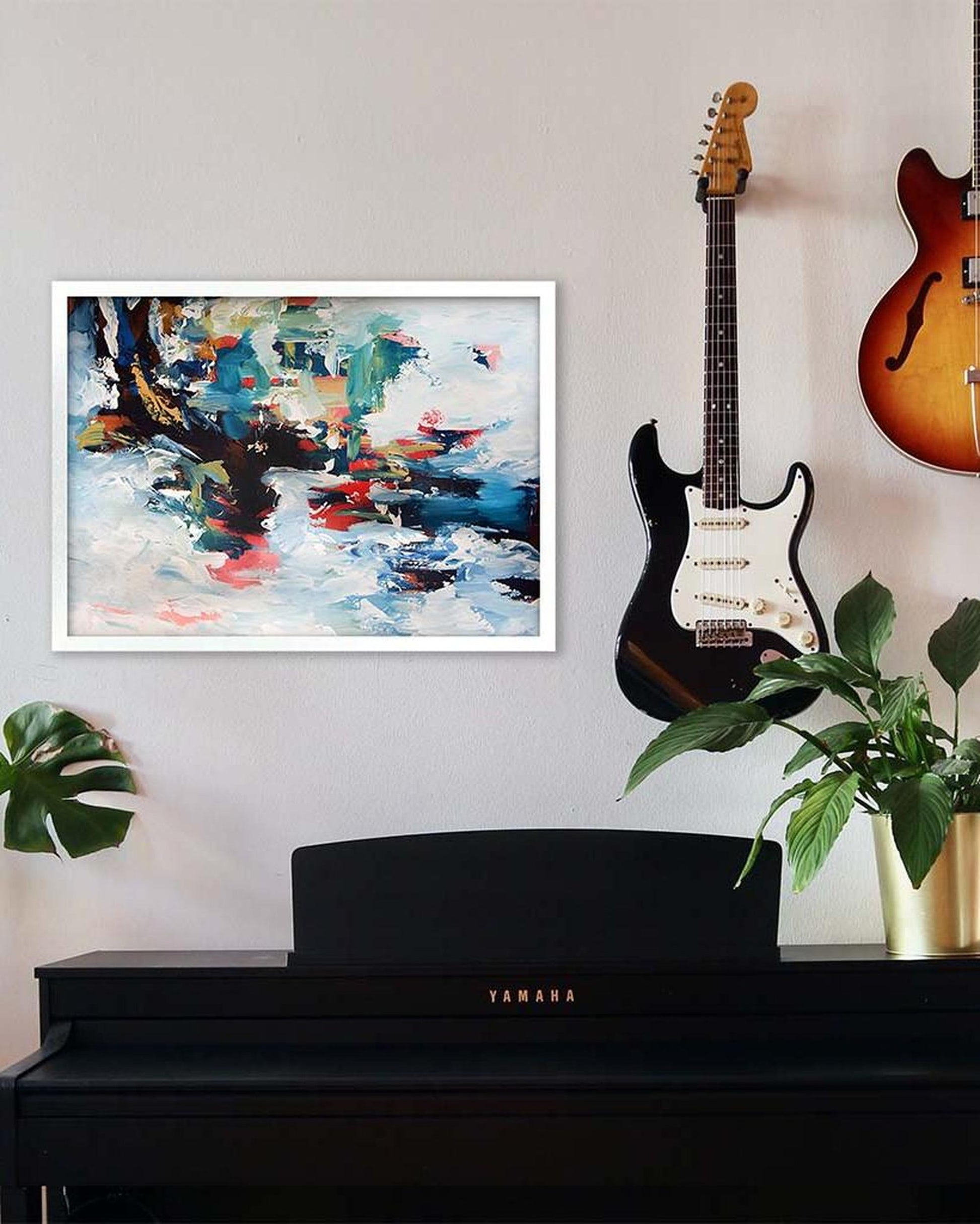 Guitar leaning against a wall next to a framed abstract painting, with a piano and plants in the foreground.