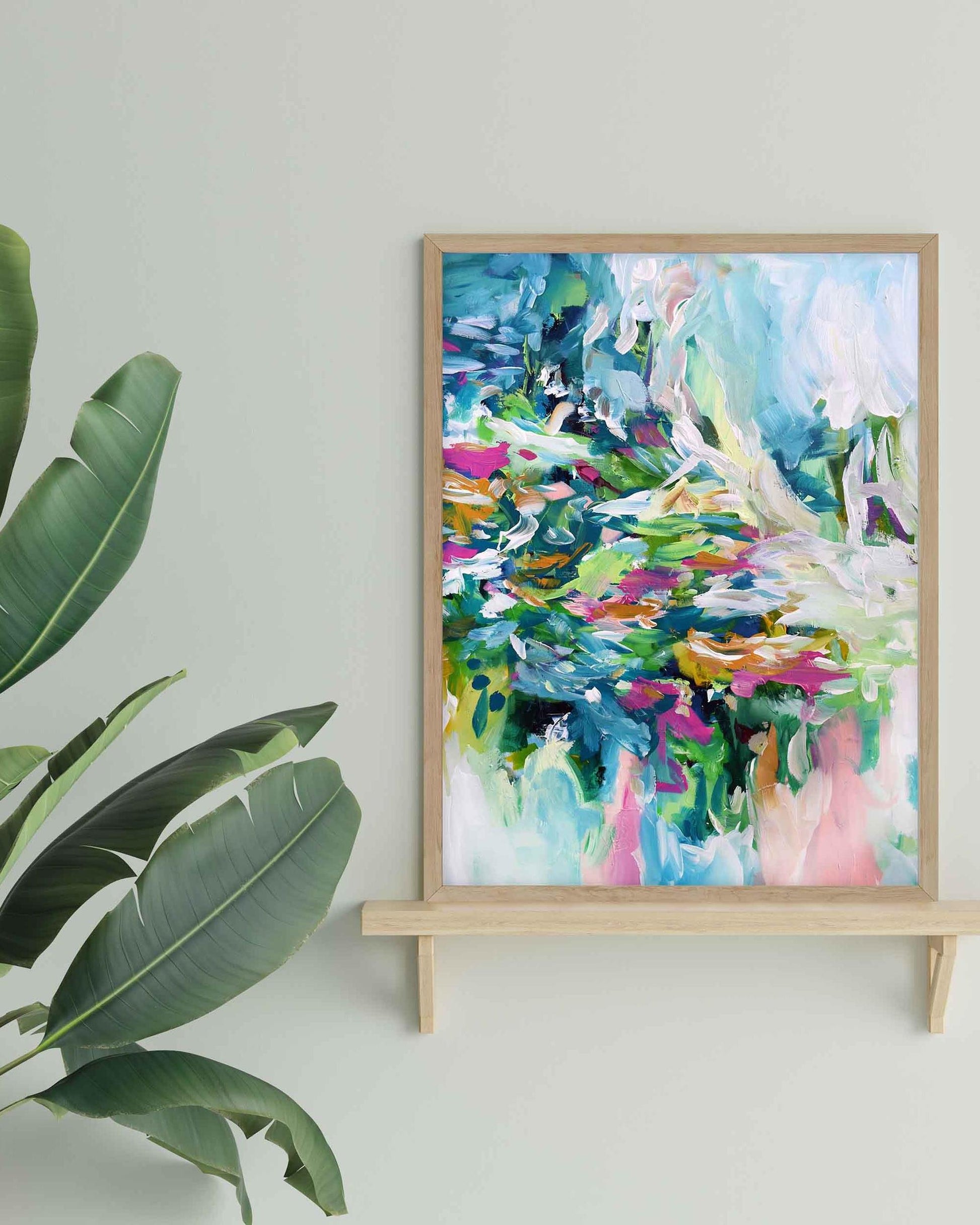 Colorful abstract painting in a wooden frame on a white wall with a plant to the left.