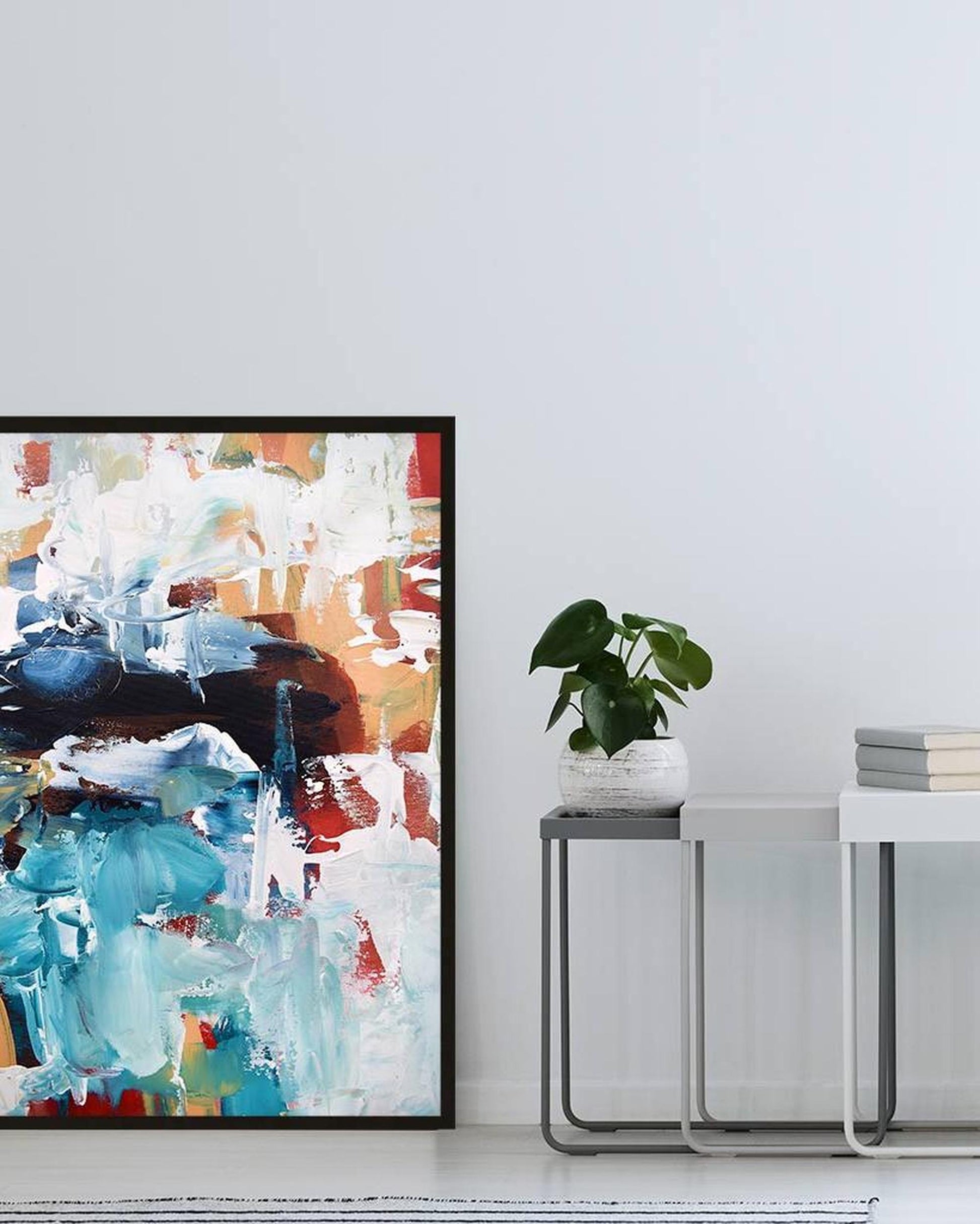 Abstract art print in a black frame on a white wall with a plant and books below.