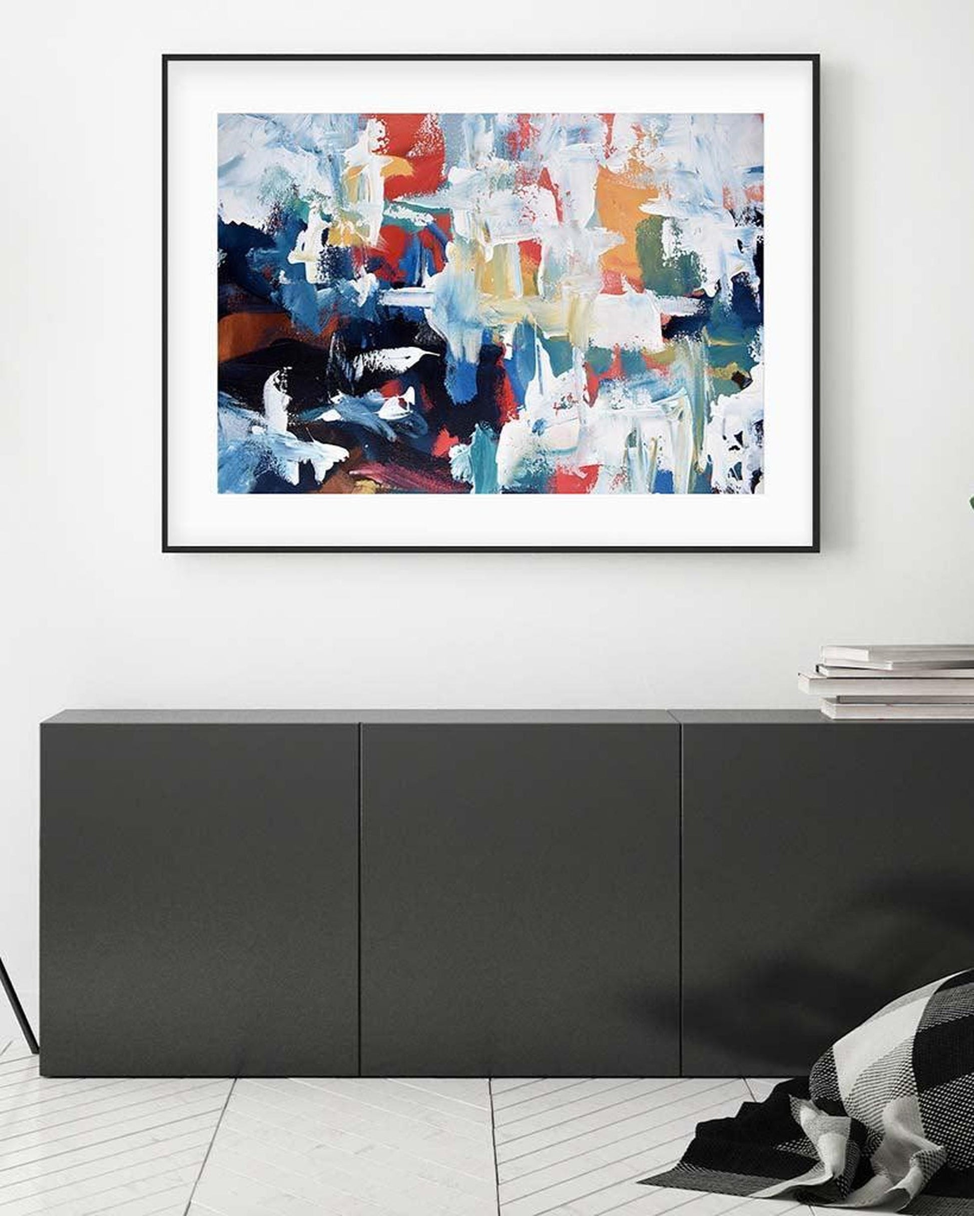 Abstract art print in a black frame on a white wall above a dark piece of furniture.