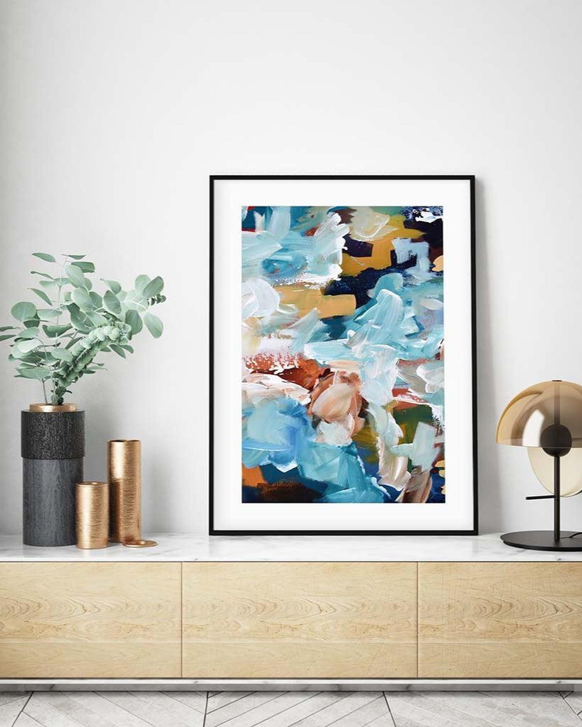 Abstract art print in a black frame on a wall above a wooden cabinet with decorative items.