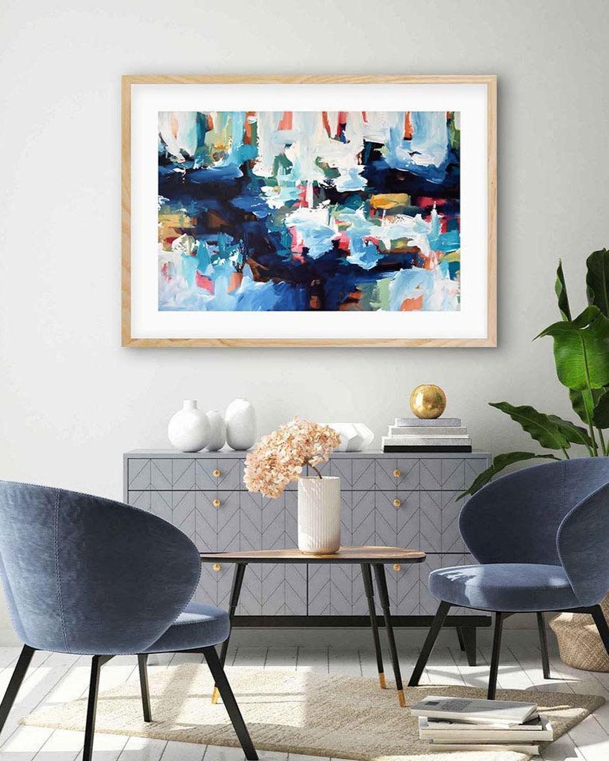 Framed abstract artwork featuring overlapping colors blue, white, yellow, and red, displayed on a wall above a grey console table with two blue chairs in front.