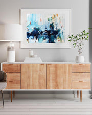 Wooden sideboard with abstract painting above, lamp to the left, and plant on the right.