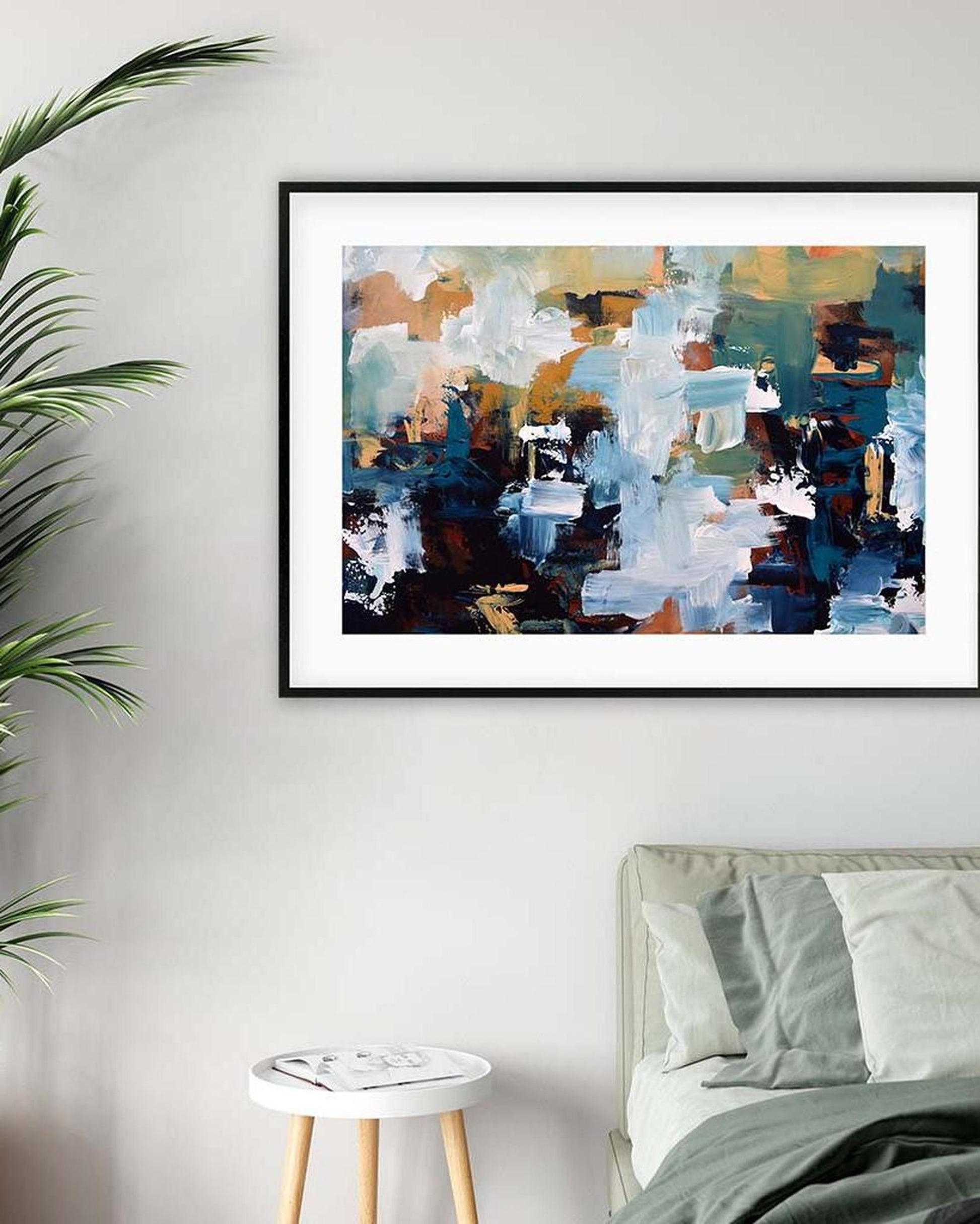 Abstract painting in a black frame on a wall above a bed with a plant to the left.