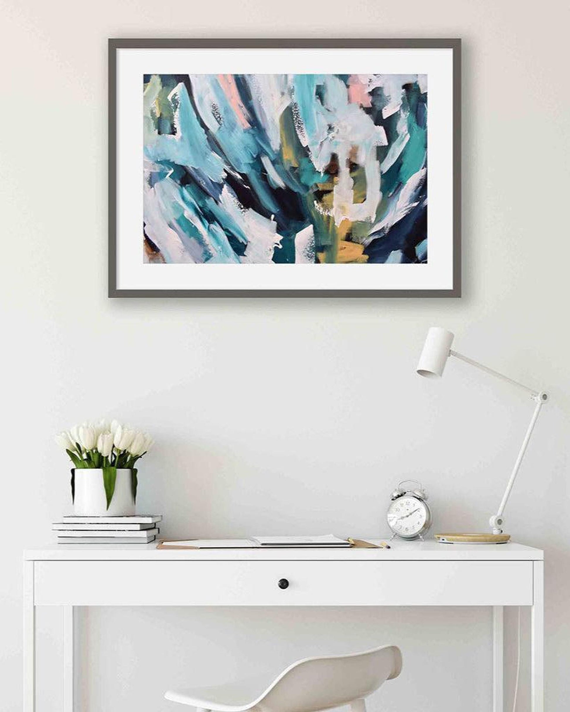 Framed abstract art above a white desk with decor items.