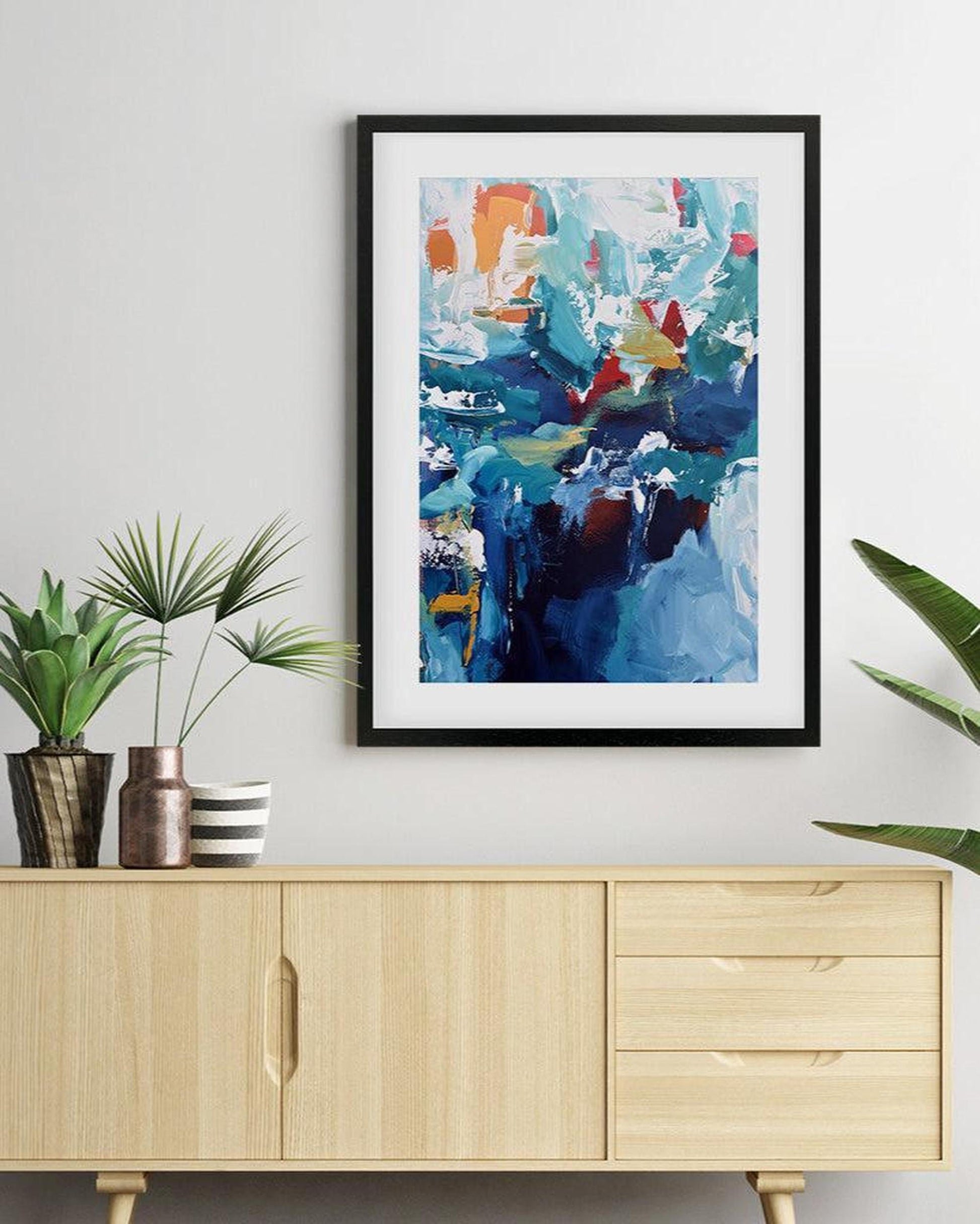 Framed abstract art piece on a wall above a wooden cabinet with plants.