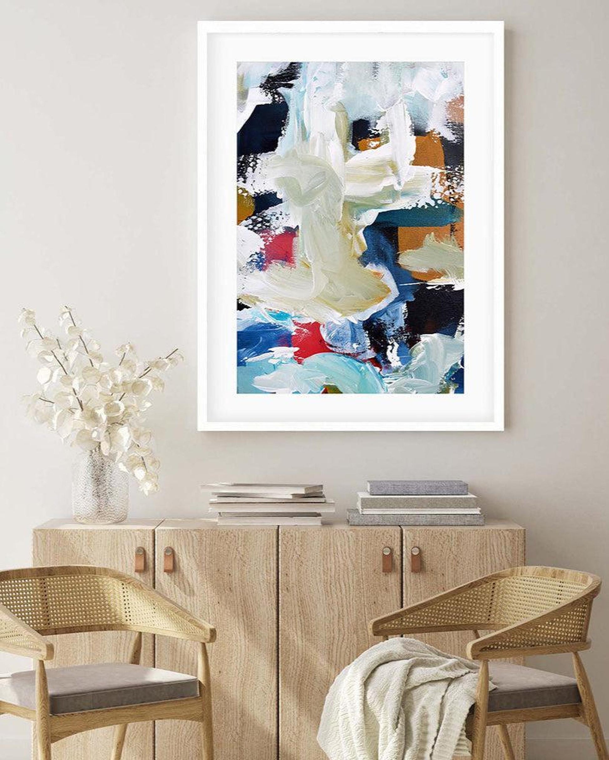 Abstract art print in a white frame on a wall above a wooden cabinet with chairs.