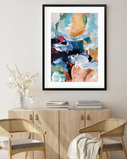Abstract art print in a black frame on a wall above a wooden cabinet with chairs.
