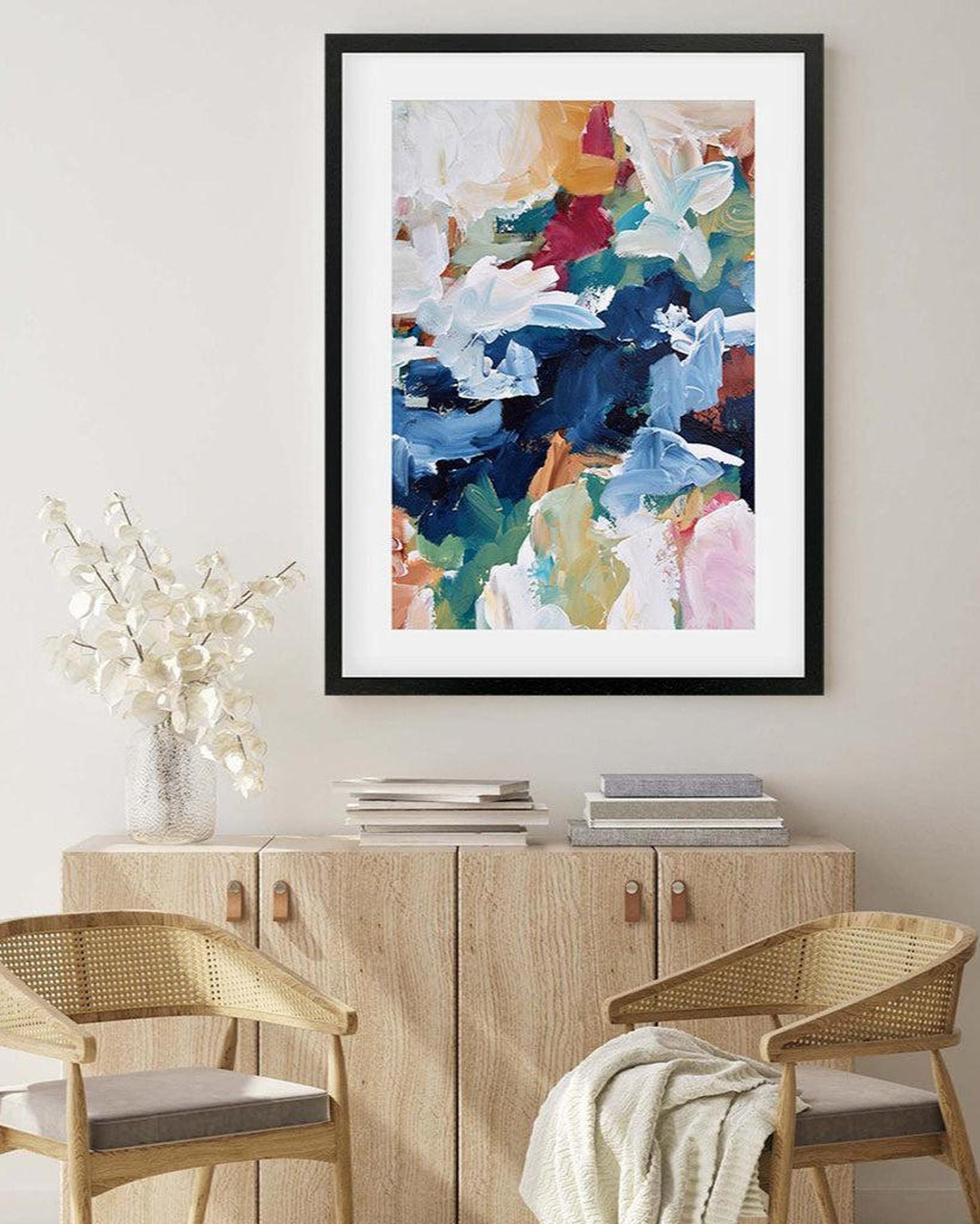 Framed abstract art piece on a wall above a wooden cabinet with chairs.
