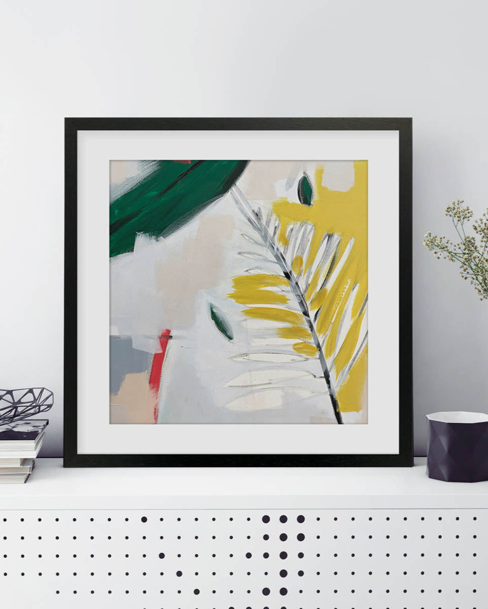 Framed abstract art piece on a white wall with a plant and books in the background