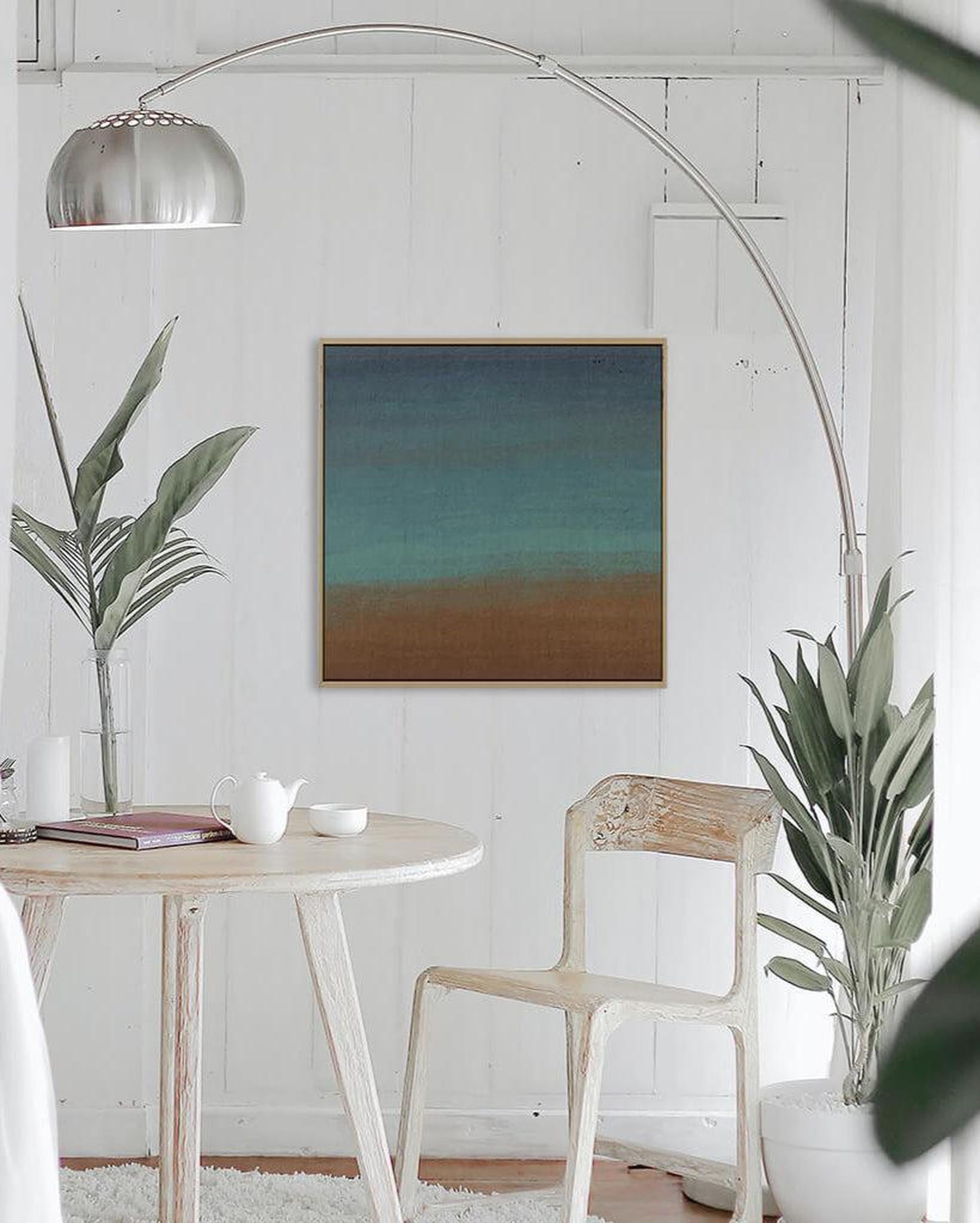 A canvas print featuring abstract geometric patterns in brown, green, and teal, framed and displayed above a wooden table with a plant and a cup, indicating a home setting.