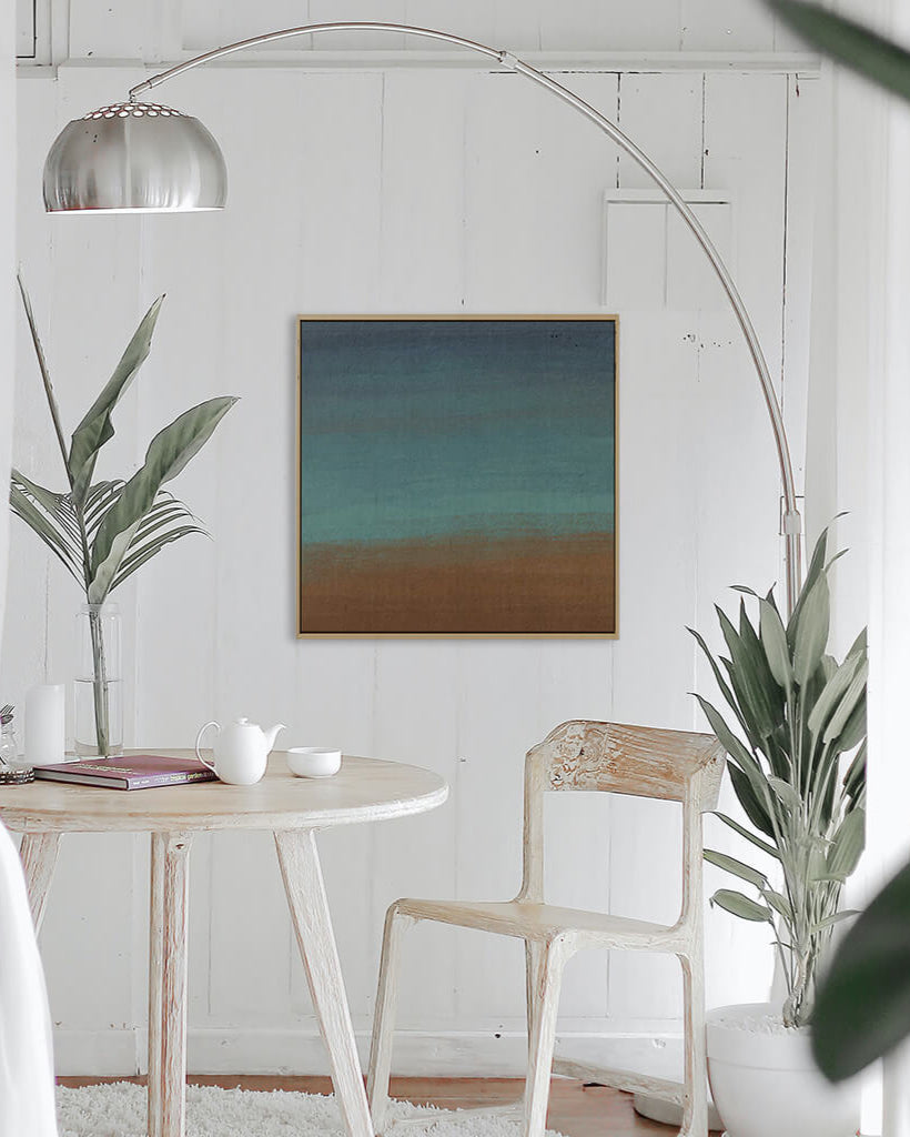 A canvas print featuring abstract geometric patterns in brown, green, and teal, framed and displayed above a wooden table with a plant and a cup, indicating a home setting.