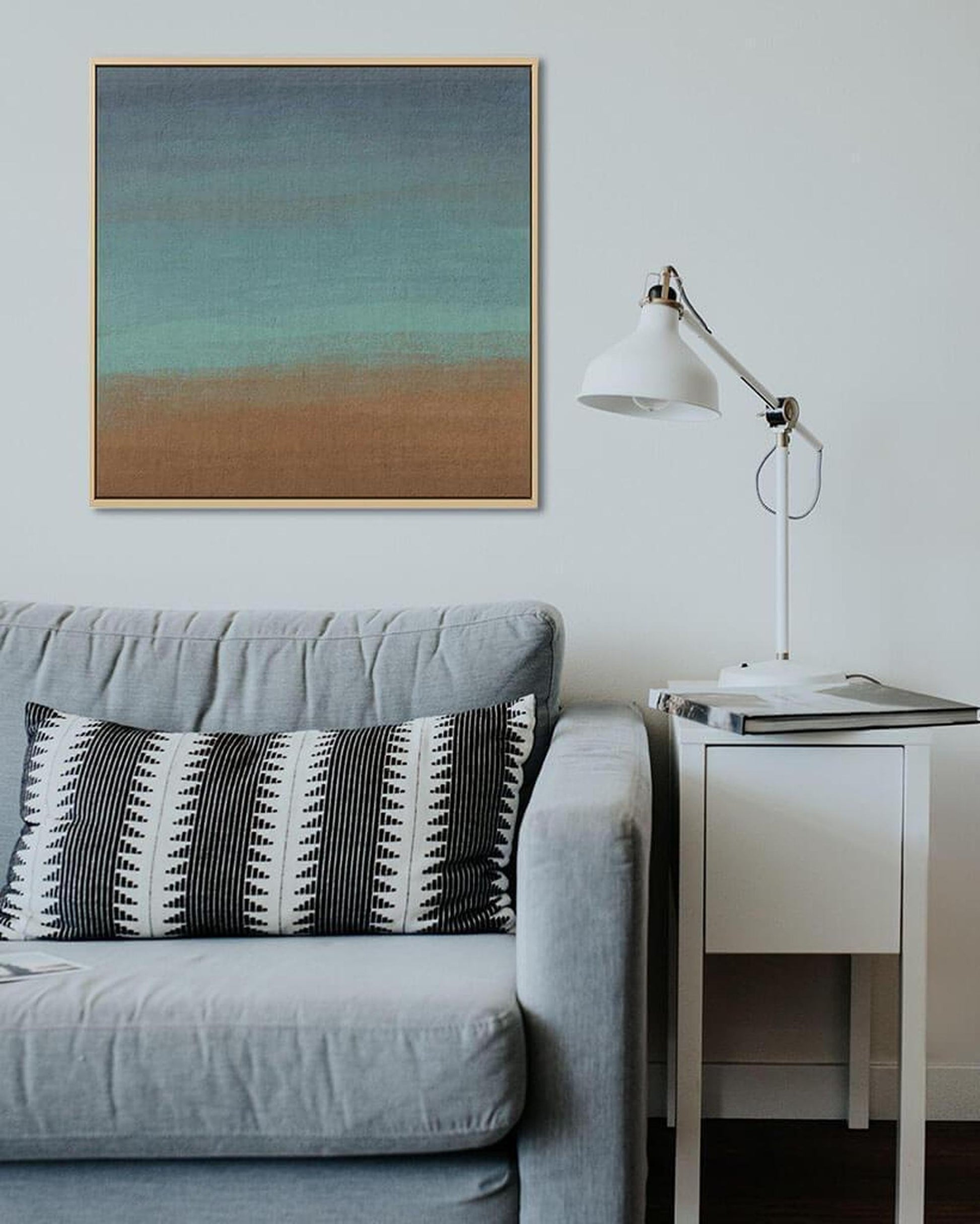 Gray sofa with a patterned pillow, white side table with lamp, and abstract painting on a light gray wall.