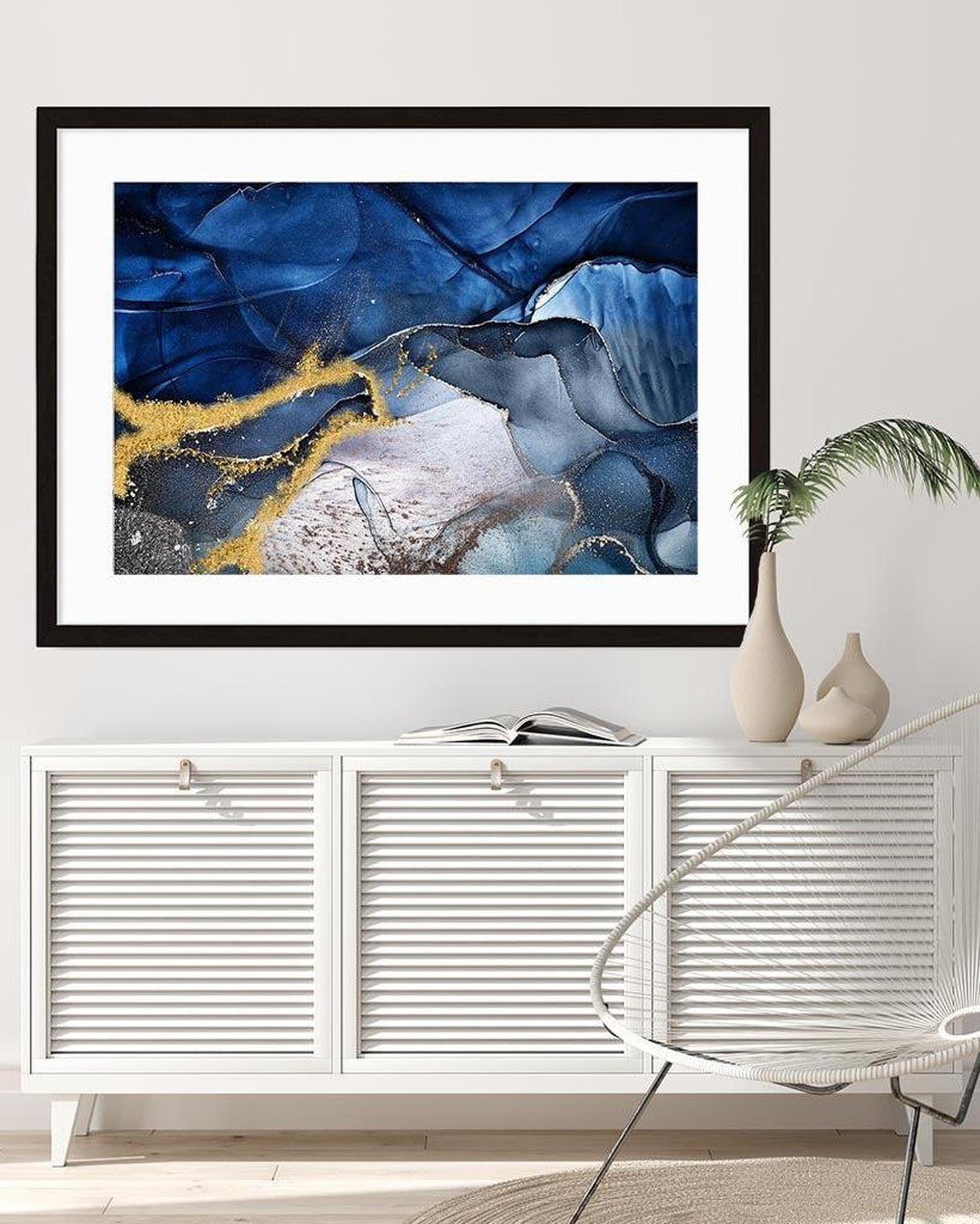 Framed abstract art piece with blue and gold tones on a wall above a white cabinet.