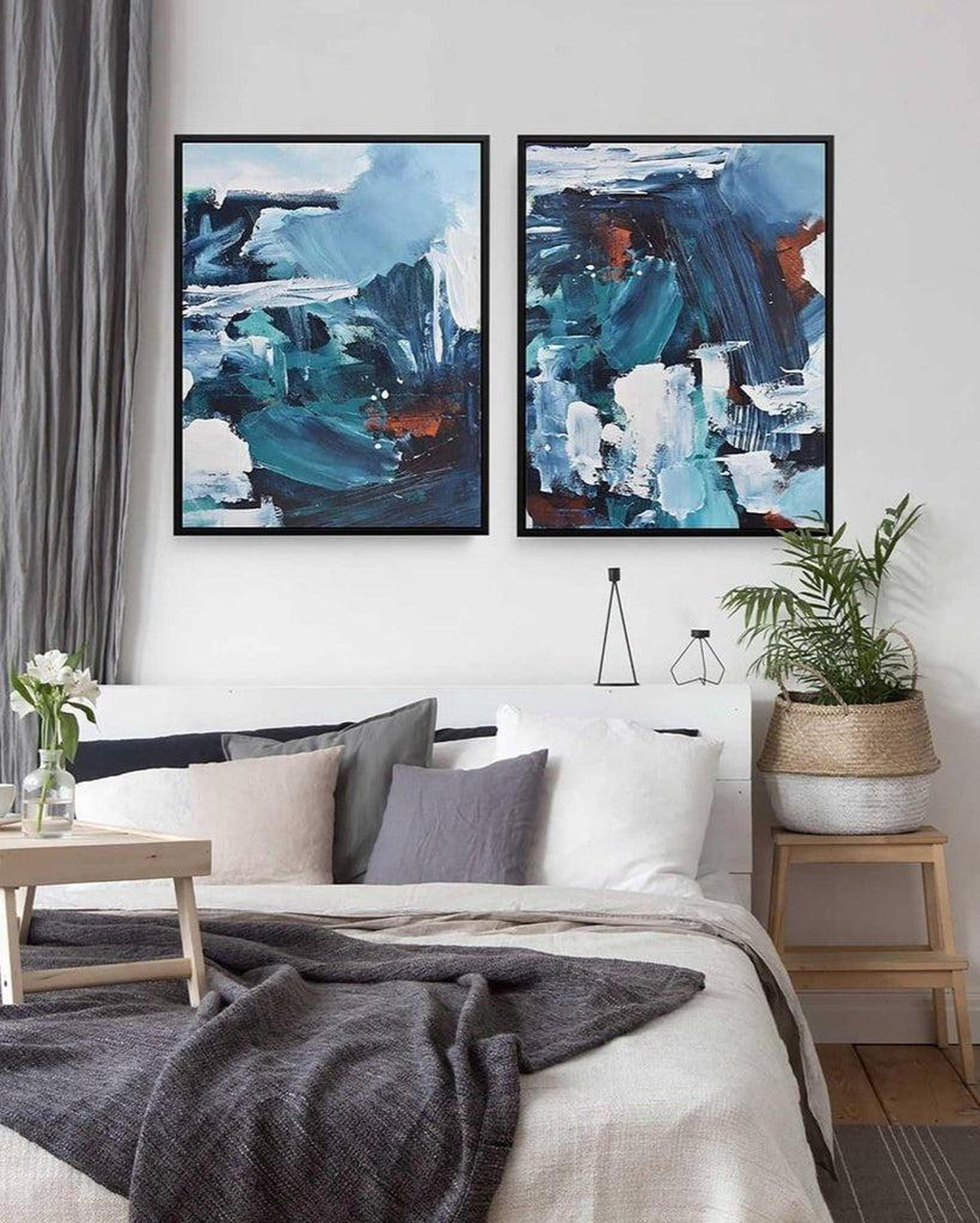 A set of two abstract canvas art pieces displayed over a white bed in a bedroom setting, with a variety of colors including blues and whites.