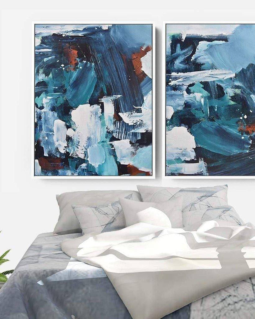 Two abstract paintings above a bed with gray bedding and white pillows.