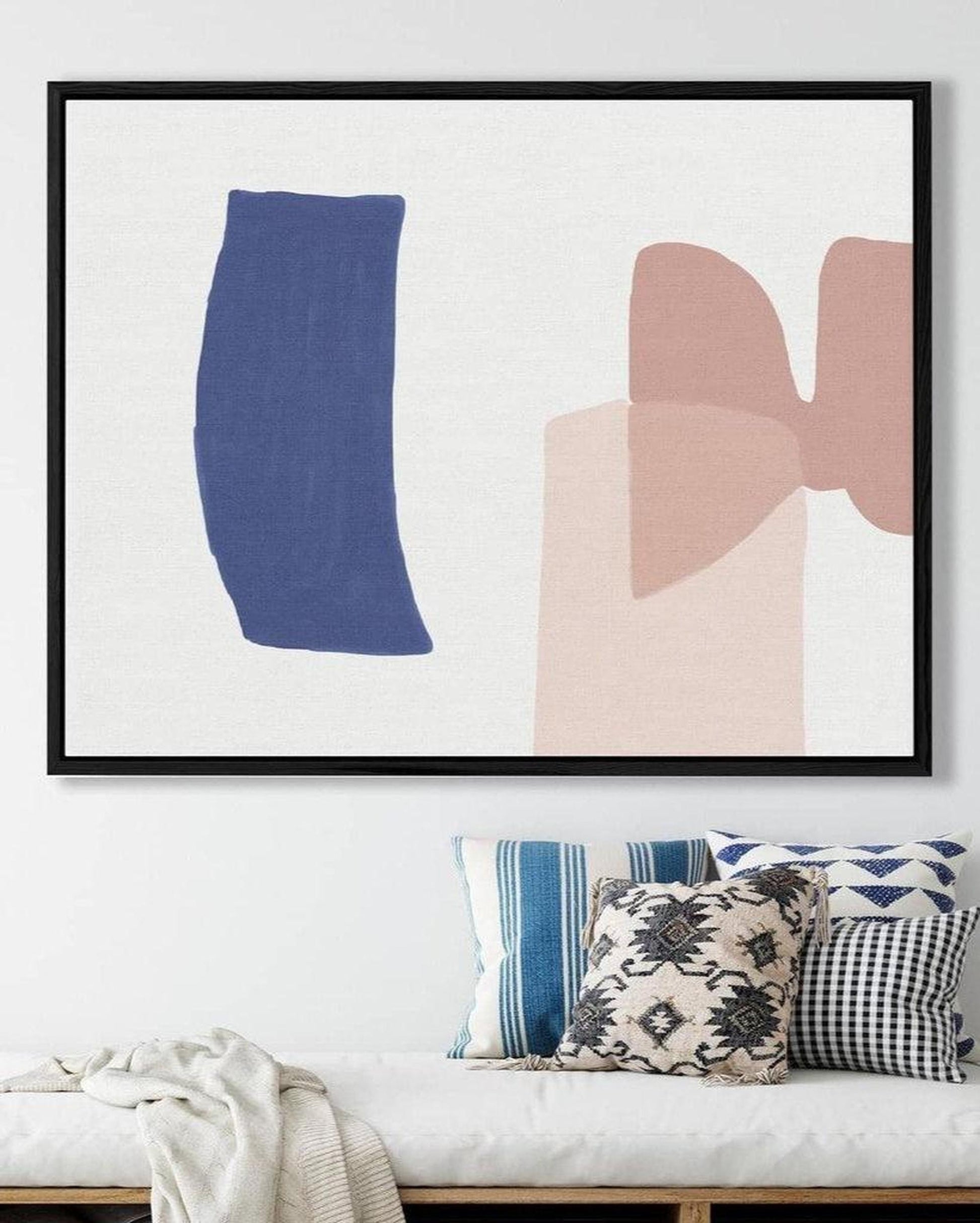 Framed abstract art print with blue and pink shapes on a white wall above a bench with patterned pillows.